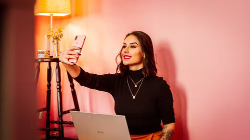Woman taking a selfie on a mobile phone in front of a pink background and next to a stool with a lamp.