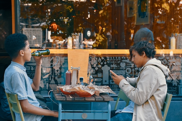 Two boys eating a meal outside, with one boy browsing on his phone.
