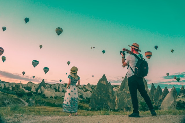 A man taking photos of a woman in front of a mountain landscape and hot air balloons.