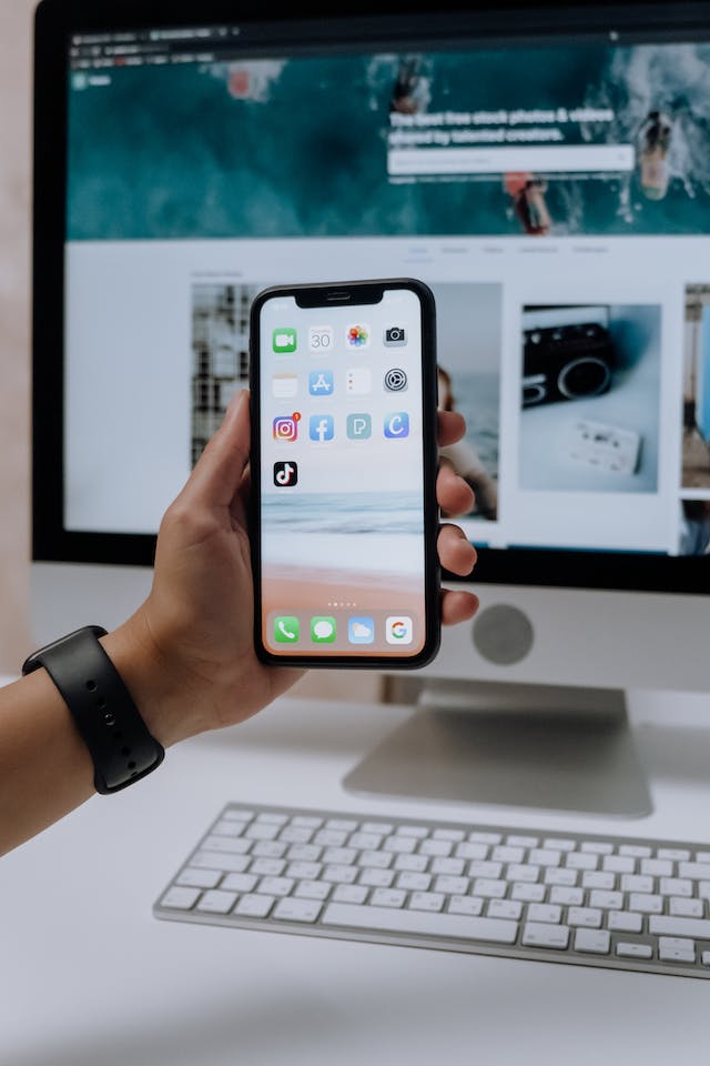 A picture of someone holding a smartphone displaying the TikTok app and other mobile applications in front of an iMac.