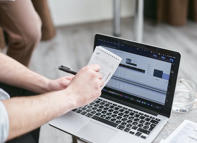 A person looks at graphs on a laptop while doing calculations on paper.