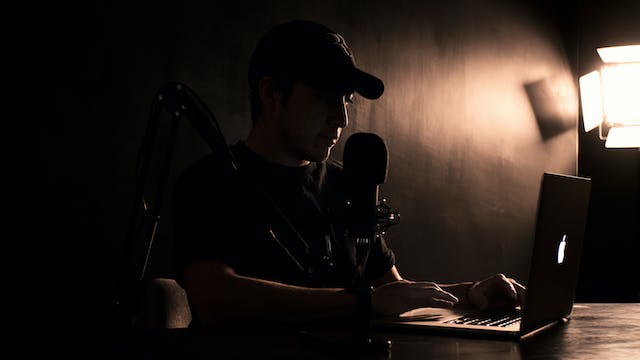 A man sits in front of a laptop and microphone for a podcast.