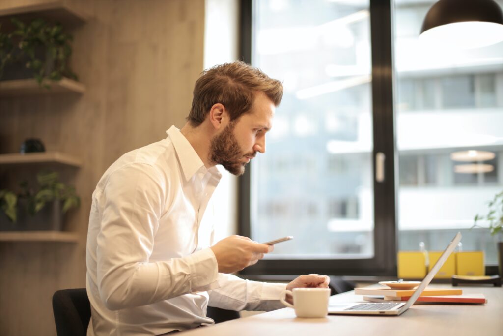 A young professional holds his phone while checking his laptop.