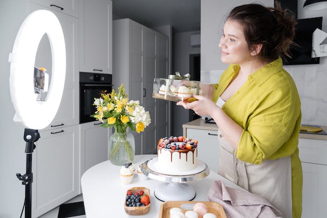 A woman in a kitchen stands in front of a ring light and a phone camera to showcase her cakes.