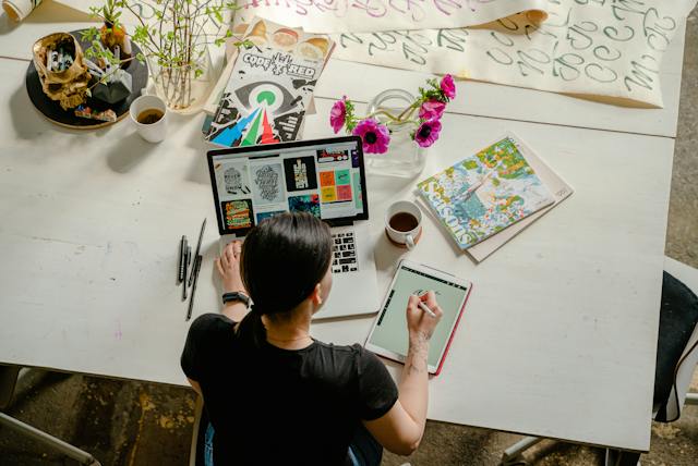 An overhead shot of a woman working on her laptop and creating digital art on a tablet.