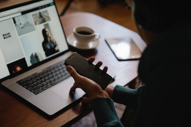 A woman browses on her laptop while holding a phone.