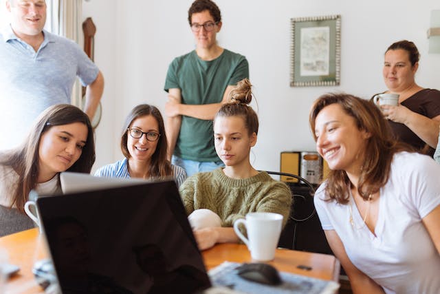 An image of a group of people sitting and standing around a table watching TikTok videos on a laptop.