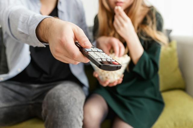 A man uses a TV remote control while sharing a bowl of popcorn with a woman.