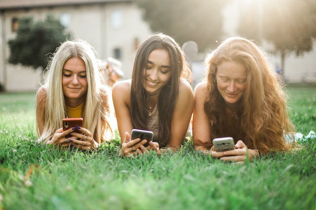Three young women lie on the grass while browsing on their phones.