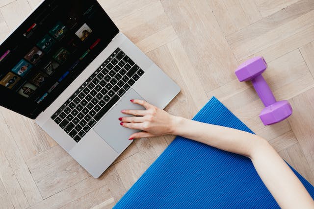 A woman on a yoga mat stretches her arm to search for TikTok exercise videos on her laptop.