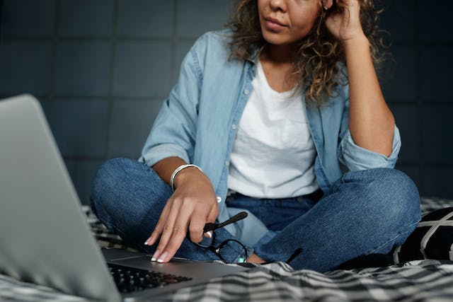 A woman sits on her bed and browses through her laptop.
