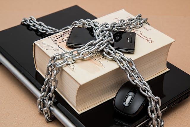 A metal chain is wrapped around a smartphone, book, and laptop.