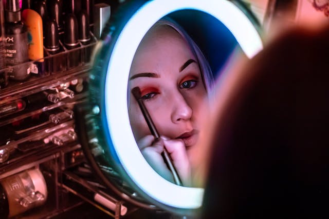 A woman sits in front of a mirror and uses a brush to apply eye shadow.