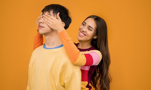 A woman stands behind a man and covers his eyes with her hands.