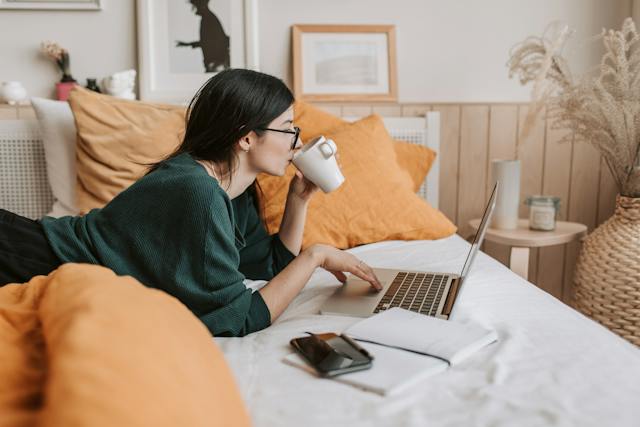 A woman is lying on her bed, drinking from a cup and browsing videos on her laptop.