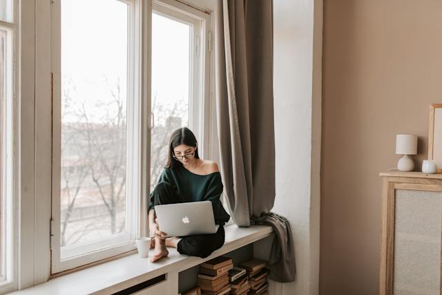 A woman sits beside her window while she watches videos on her laptop.