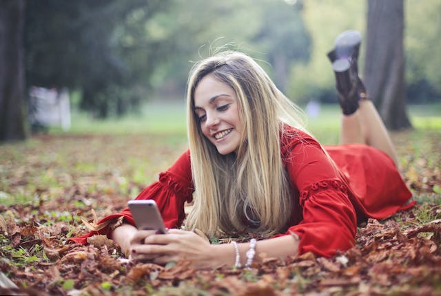 A woman lies on her stomach on fallen leaves as she scrolls on her phone.
