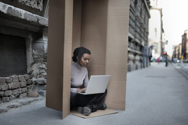 A woman sits inside a large cardboard box while using her laptop.