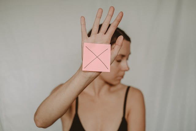 A woman holds up a square piece of paper with an X on it.