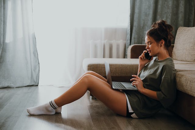 A woman sits on the floor while talking on the phone and browsing on her laptop.