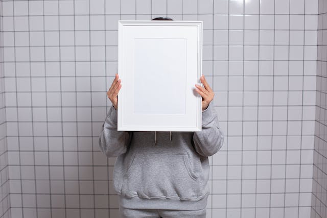 A person stands in a white, tiled room and hides behind an empty, white picture frame.