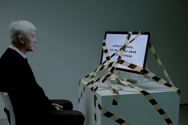 An older man sits in front of a computer monitor covered in black and yellow caution tape.