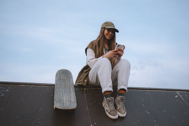 A young woman rests beside her skateboard and watches TikToks on her phone.