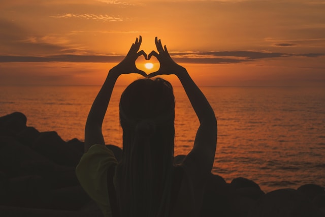 A woman’s silhouette frames the setting sun inside her heart-shaped fingers.