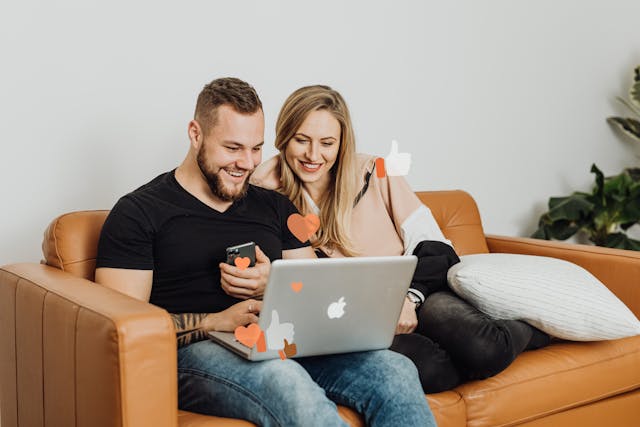A couple on a couch smiles as they check out their laptop.
