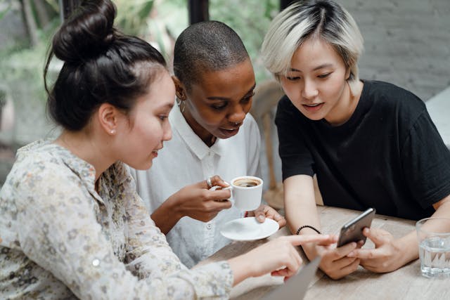 Three female friends sit at an outdoor cafe and use a phone to check social media updates.