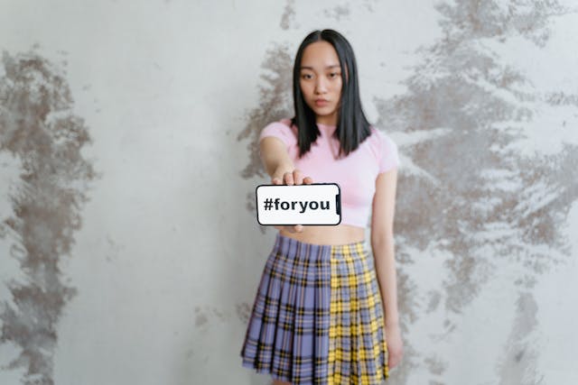 A young woman holds up a phone that shows the hashtag “#foryou” on its screen.