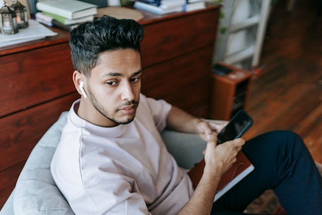 A man sits in a chair with his phone in his hand and an Airpod in his ear.