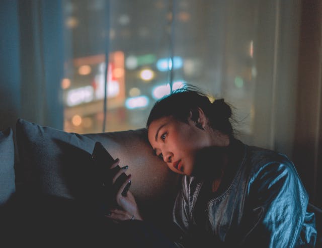 A woman in a dark bedroom puts her head on a pillow and checks her phone.