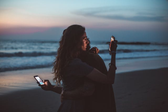 A couple at the beach hugs each other while they also look at their phones.