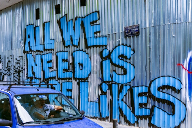 The side of a building shows a spray-painted sign that says, “All we need is more likes.”