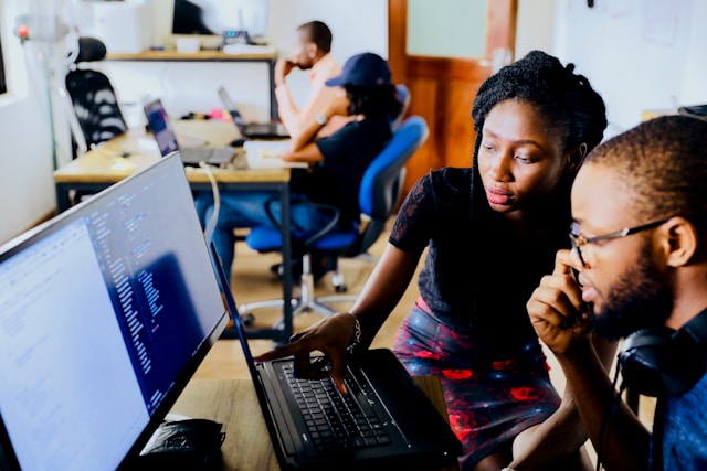 Two people sit in front of a computer that displays computer code elements.