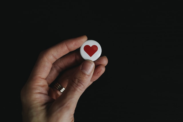 A hand holds a tiny white button with a red heart on it.