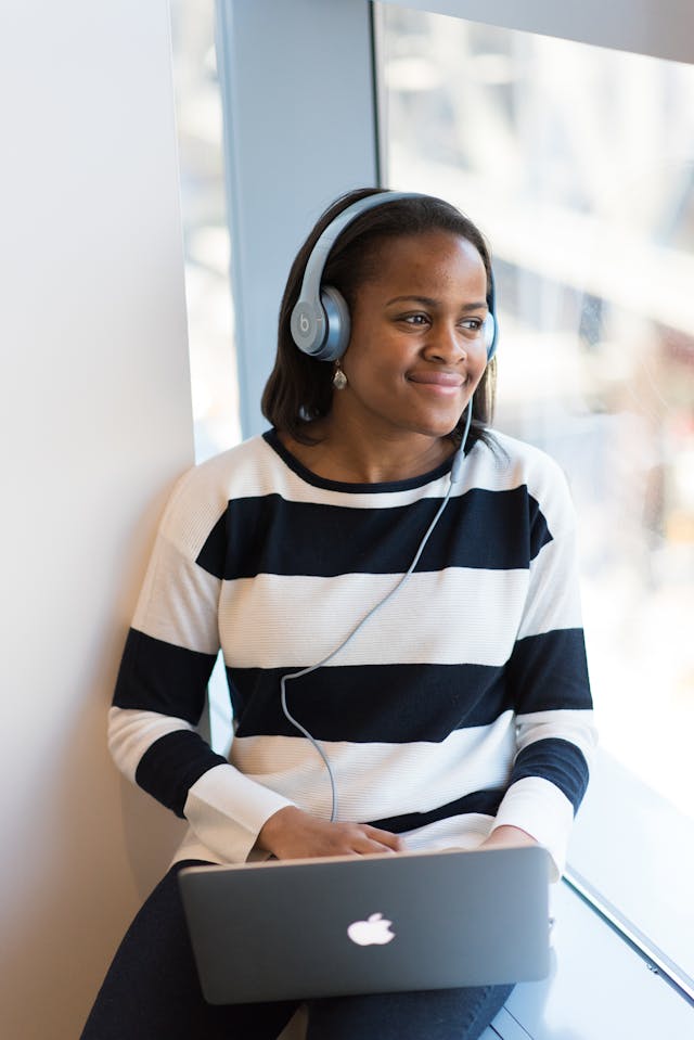 A woman in a striped shirt listens to headphones connected to her laptop.
