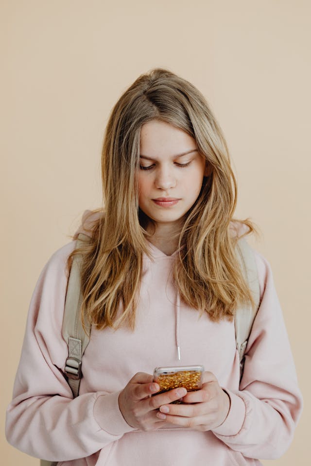 A girl in a pink hoodie scrolls on her phone.