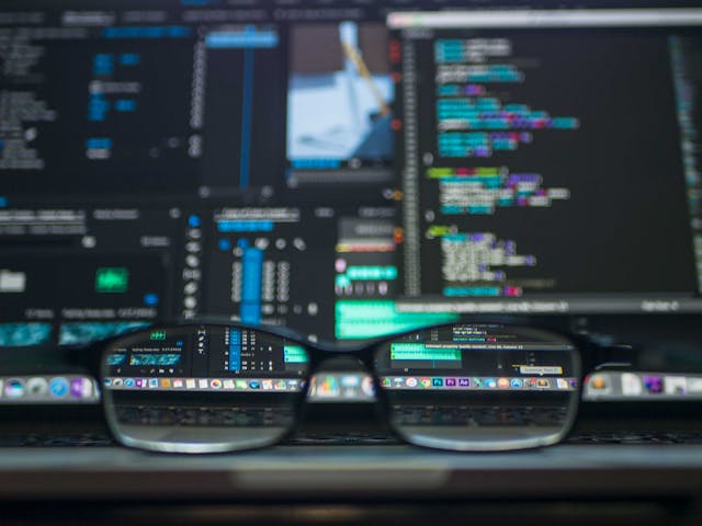 A pair of prescription glasses in front of computer monitors displaying coding elements.