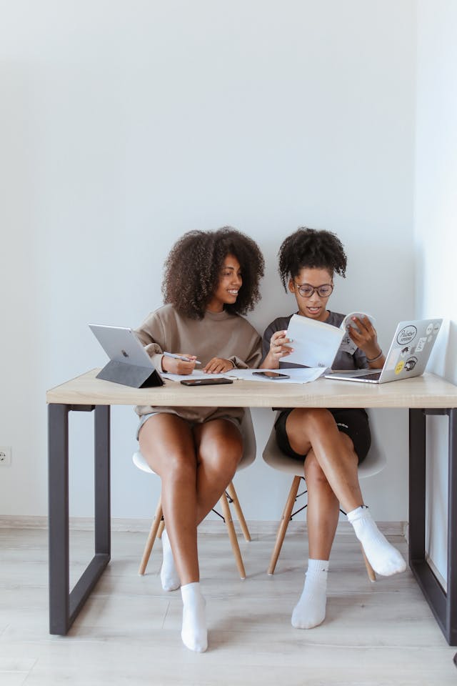 Two women sit at a desk with their laptops.