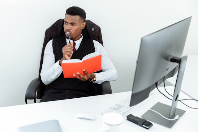 A man sits in his office in front of his computer and holds a notebook and pen.