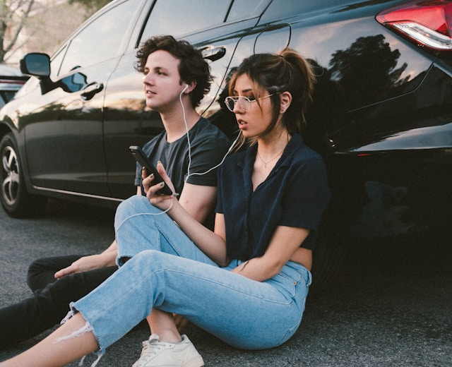 A man and a woman lean against a car and share earphones to listen to music on a phone.
