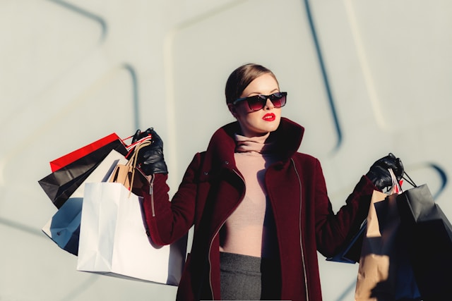A woman in a dark red coat and sunglasses carries shopping bags with both hands.