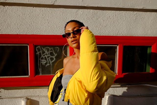 A model wears hoop earrings, sunglasses, and a bright yellow jacket over a gray tube top.
