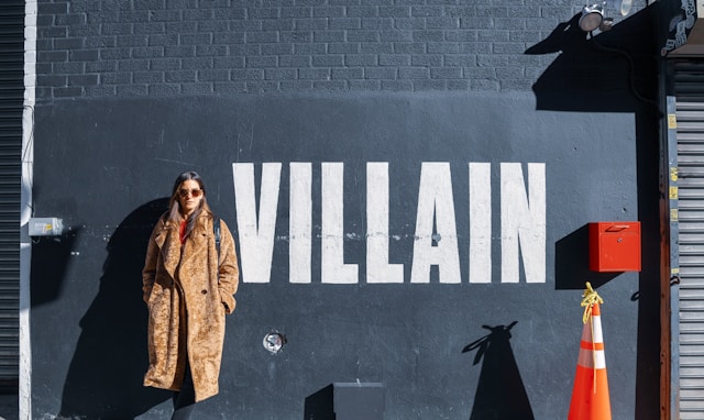 A woman in a brown fur coat stands against a wall with the word “Villain” painted on it.