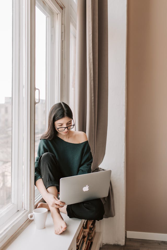 A woman sits on her windowsill with her laptop.