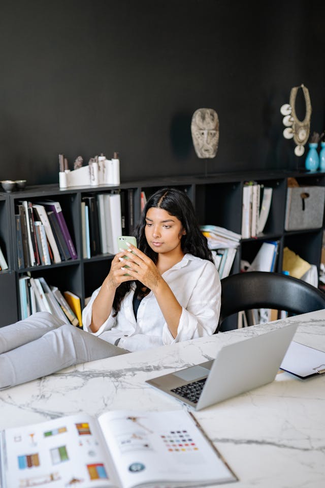 A woman scrolls on her phone while sitting in front of her laptop.