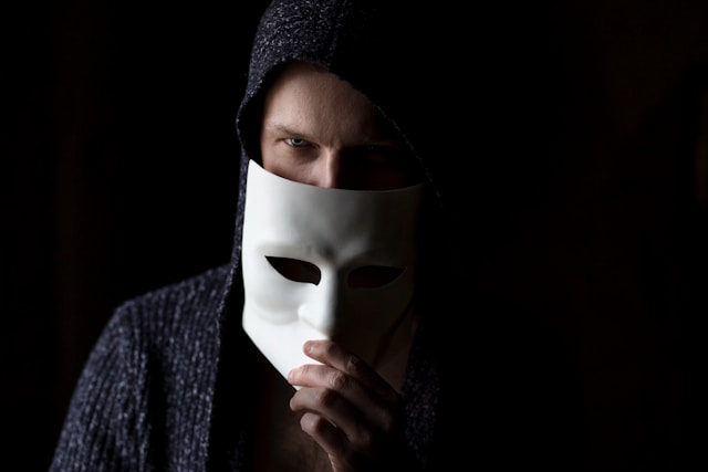 A person in shadow hides half their face behind a white mask.