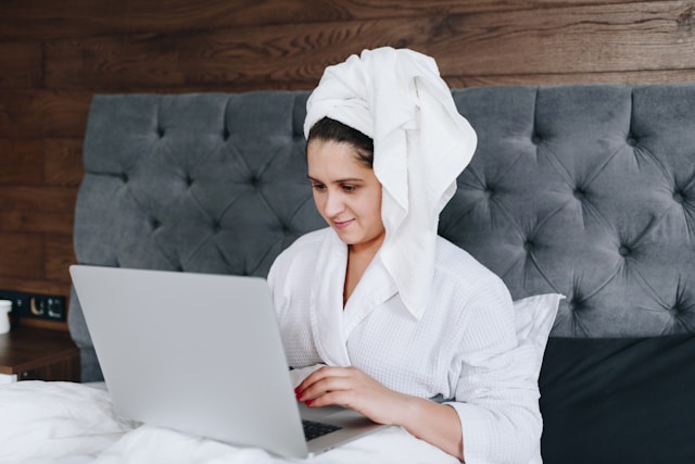 A woman wears a towel around her head and a robe and sits on the bed while browsing her laptop.